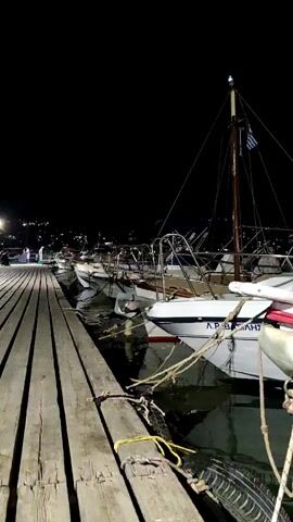 A vertical video shows a nighttime view of a harbor with several boats docked along a wooden pier. The pier takes up the left side of the frame, with weathered wooden planks stretching into the distance. Several boats are moored alongside the pier, their white hulls reflecting in the dark water. One boat in the foreground has red trim and a name painted on its side in Greek letters, "ΑΡ. ΒΑΣΙΛΗΣ". The boats are silhouetted against the dark night sky, with a few lights visible in the distance. A mast with a Greek flag is visible on one of the boats. There are no people clearly visible in the immediate foreground. The overall impression is a quiet, peaceful scene at a harbor after dark.
