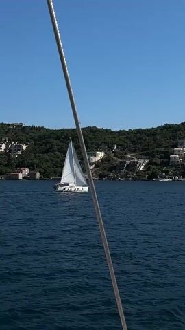 The video shows a sailboat on the water on a clear, sunny day. The camera is partially obstructed by a thick, gray rope, suggesting it is on a different boat. In the background, there is a tree-covered hill dotted with houses and a clear blue sky. The water is a deep blue, with some small waves.