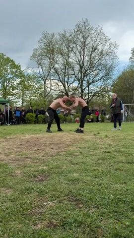 Two men with bare chests, wearing black pants and athletic shoes, are engaged in a physical altercation on a grassy field. They are grappling with each other. A crowd of spectators, some seated and some standing, surrounds the edge of the field, watching the fight. In the background, there are trees, a soccer goal, and a man in a vest and slacks walking to the side. The sky is overcast. The grass on the field is patchy, with areas of bare earth.