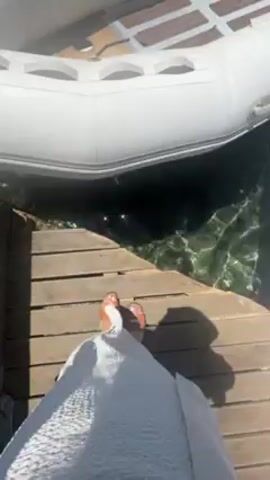Here is a concise description of the video:

The video shows a person walking from a wooden dock onto a white inflatable boat. The boat is docked near other similar boats in a clear, turquoise body of water. In the distance, a hilly coastline is visible under a clear, blue sky.  A man is seen on the inflatable boat, attending to something near the front. The video appears to be taken on a sunny day.