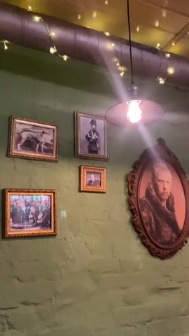 The video displays an interior shot of a room, likely a pub or restaurant, characterized by a textured green wall adorned with various framed pictures. A prominent light fixture with a glowing, exposed bulb hangs from the ceiling, casting a bright glare on the scene. Above the framed pictures, a large industrial-style pipe or duct runs horizontally, decorated with small, warm-toned string lights.

Several framed photographs are mounted on the green wall. Directly below the main light source is a small, gold-framed black and white portrait of a man in a suit and bow tie, identifiable as Winston Churchill. Above it hangs a larger, gold-framed black and white portrait of a man in a military uniform with a distinctive tall, feathered hat and medals. To the right, a large, ornate oval frame holds a sepia-toned portrait of a bearded man wearing a fur-lined coat. On the left side of the wall, there are two more gold-framed pictures: one featuring an animal (possibly a wolf or large dog) in a sepia tone, and another, smaller one depicting a group of people.

The ambiance suggests an evening setting, illuminated by artificial light. Background chatter and laughter in a foreign language (Russian) can be heard, indicating a social environment. The overall decor combines rustic elements with historical portraits. No discernible text is visible within the frames or on the wall. The location is contextualized as Заволжье, Russia.
