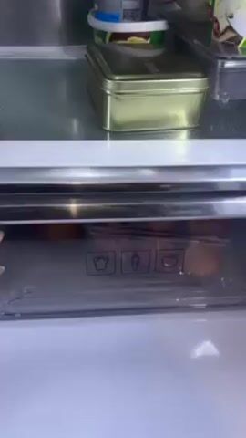 The video shows a refrigerator drawer being opened to reveal fruit. The drawer contains mangoes, apples, and a package of nectarines. Above the drawer are items on a shelf, including a container of butter, a rectangular gold-colored tin, and a plastic container with food. The refrigerator interior is white and metallic.