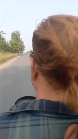A person with reddish-brown hair is riding a motorcycle on a paved road. The camera is positioned behind the rider, capturing their back and the road ahead. The rider wears a plaid shirt and sunglasses. The road is flanked by trees and dry vegetation on either side. In the distance, a white car and a large truck are visible on the road ahead. The sky is clear and bright.