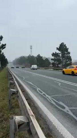 The video shows a multi-lane road on a gray, overcast day, likely a highway. Several vehicles are traveling on the road, including a yellow car in the foreground, followed by a silver SUV, a white van, and other cars further down the road. The camera is positioned at the side of the road, behind a metal guardrail. Trees line the roadside, and a cell tower is visible in the distance. The road markings are faded and worn. There are no people visible in the video. The cars move from right to left in the frame. The general tone of the video is unremarkable and ordinary.
