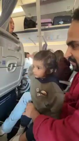 Inside an airplane cabin, a young child sits on a man's lap, both facing forward. The child, wearing light-wash jeans and a green hooded sweatshirt with decorative patches, looks up and to the side. The man, wearing a red shirt, holds the child. The child reaches towards the closed tray table attached to the seat in front. Other passengers are visible in the background, along with overhead luggage compartments filled with bags. The cabin interior is typical of a commercial airplane.