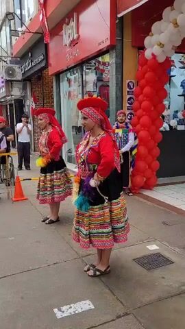 Two women in traditional red and colorful Andean attire, including headdresses, skirts, and embroidered tops, stand on a sidewalk in front of a store. The woman in the foreground holds a bouquet of colorful feathers and moves her arms. The woman behind her mirrors this motion. A man in a colorful outfit with a feathered hat stands to the right. The store has a red awning with white lettering. Red and white balloons are attached to the store's facade. Other people are visible in the background. An orange traffic cone is on the sidewalk.