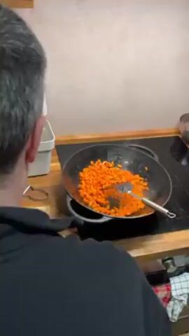 A gray-haired man wearing a black sweatshirt is cooking in a kitchen. The man is facing away from the camera, stirring diced carrots in a large black pan on a glass-top stovetop. Steam is rising from the pan. A black shelf is visible above the stove. A few containers and a metal bowl are on the counter to the right of the stove. The countertop is made of wood.