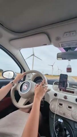 The video shows a person driving a car. The driver's hands are on the steering wheel, and the car's interior is visible, including the dashboard, phone mount, and gear shift. Outside the car, a road can be seen stretching into the distance, with a field of wind turbines on either side. The sky is cloudy.
