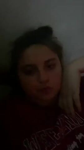 A blurry video shows a young person lying on their side, facing the camera. Their hair is pulled up into a bun. They are wearing a dark-colored shirt. Their right arm is bent and resting near their head. The background is dark and appears to be a wall. The lighting is dim.