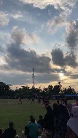 A low-angle, vertical shot captures a soccer match on a grassy field as dusk approaches. The sky is filled with dramatic clouds, with the setting sun peeking through on the right side of the frame. A tall cell tower stands in the distance. On the field, several players in red uniforms are actively engaged in the game, some near the center and others towards the left. Spectators, mostly women wearing hijabs and some men, are seated or kneeling on the grass in the foreground, watching the match. The overall atmosphere suggests a casual, community sporting event during twilight.