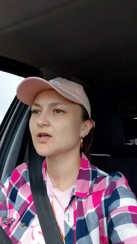 The video shows a woman in a car. She is wearing a pink baseball cap, a pink and blue plaid shirt over a pink t-shirt, and a seatbelt. She is looking to her left and speaking. The interior of the car is visible, including the dashboard and the passenger seat. The background outside the car appears to be trees and foliage.
