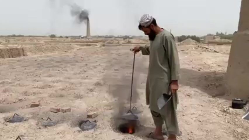 A man stands in a dusty, barren landscape near a brick kiln that is emitting black smoke. He wears a long, light-green robe and a white head covering. In one hand, he holds a long metal tool attached to a flat, dome-shaped object above a fire burning in a hole in the ground. The other hand holds a small metal scoop. Several similar dome-covered fires are burning nearby. A stack of bricks sits to the left, and a brick structure is visible to the right. A metal kettle sits on the ground next to the brick structure. Piles of bricks are visible in the distance.