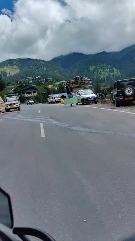 The video shows a white car on a paved road in a mountainous area. The car has its passenger door open, and a person is partially visible inside, holding a large, flat, light-colored object that is extending out of the car. The object appears to be a piece of furniture or a large panel. The car is parked on the right side of the road, which is a two-lane road. In the background, there are more cars, some buildings on a hillside, and tall green mountains with a cloudy sky. Several yellow bags are piled up on the side of the road near the car. The perspective of the video suggests it is being filmed from a moving vehicle, possibly a motorcycle, as a portion of a rearview mirror is visible in the foreground.