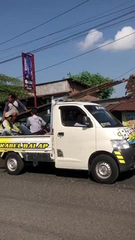 A white pickup truck is driving on a paved road under a clear blue sky with some clouds. The truck is carrying a large bundle of long, slender objects, possibly bamboo poles or pipes, tied together and extending out of the back. There are at least three people in the bed of the truck, with one sitting on top of the cargo and two others seated behind the cab. One person in the cab appears to be driving. The truck has some decorations on the front, including a sticker with a cartoon character. In the background, there are buildings, trees, and power lines. A banner is attached to a pole on the left side of the frame, displaying some text and an image. The overall scene suggests a practical, perhaps commercial, activity.