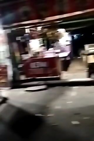 The video shows a blurry, nighttime street scene with several food stalls. In the first few seconds, a red and white sign is visible on a stall. Then, a person walks past a stall with a red and white sign and a motorcycle parked in front. Later, a person with a dark shirt and blue jeans walks towards a stall with a colorful umbrella and a bright light. The camera is shaky throughout the video, making it difficult to discern fine details.