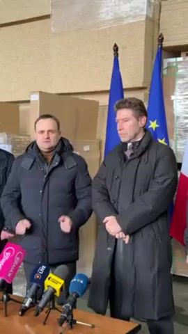 Two men are standing behind a table with microphones. Behind them are pallets of goods stacked high, as well as three flags, two European Union flags, and one Italian flag. More people are standing behind the men and flags. The men are wearing winter coats.