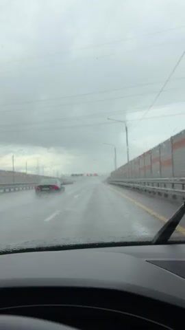 THE VIDEO SHOWS A POV SHOT FROM INSIDE A VEHICLE DRIVING ON A WET ROAD DURING RAIN. THE WINDSHIELD WIPERS ARE ACTIVATED AND WIPING THE RAIN FROM THE WINDSHIELD. THE ROAD IS A MULTI-LANE HIGHWAY WITH OTHER VEHICLES VISIBLE IN THE DISTANCE. THERE ARE CRASH BARRIERS AND SOUND BARRIERS ALONG THE SIDE OF THE ROAD. THE SKY IS OVERCAST AND GRAY, INDICATING POOR WEATHER CONDITIONS. THE VIDEO CAPTURES THE EXPERIENCE OF DRIVING IN RAIN.