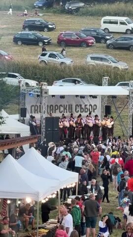 A outdoor festival scene with a stage set up for a performance. A group of musicians dressed in traditional clothing are playing instruments on the stage. A large crowd of people are gathered in front of the stage, watching the performance. There are several white tents set up around the area, likely for vendors or shade. In the background, a grassy hillside is visible with several parked cars. The overall atmosphere appears to be a lively community event.