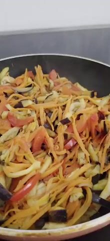 A close-up shot shows a black frying pan filled with chopped vegetables being stir-fried. The vegetables appear to be carrots, eggplant, and tomatoes, all cut into thin strips. The camera is positioned directly above the pan, and the lighting is somewhat dim, suggesting it might be indoors. There is a slight haze rising from the pan, indicating that the food is hot. The background is blurred, but it seems to be a kitchen countertop with a dark backsplash. The video appears to be a short clip, possibly part of a cooking demonstration or a recipe video. The overall impression is of a colorful and healthy dish being prepared.