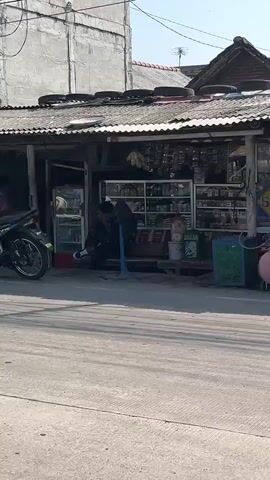 The video shows a street scene in front of a small shop. A person is kneeling on the ground in front of the shop. A motorcycle is parked to the left of the shop. The shop has shelves stocked with various items. The roof of the shop is made of corrugated metal and has several tires placed on it. The environment appears to be an outdoor commercial area.