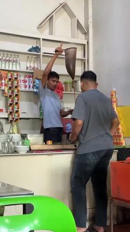 A man in a light blue t-shirt is pouring a brown liquid from a large, conical filter bag held above his head into a cup held by another man in a dark grey t-shirt. The man in the blue shirt is standing behind a counter with shelves stacked with bottles, cans, and other items. The man in the grey shirt is facing away from the camera, looking down at the cup. The background shows a wall with shelving units filled with various products. The lighting is dim, suggesting an indoor environment.