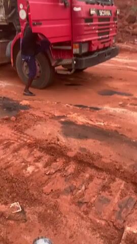 A red dump truck is parked on a dirt road. The truck is facing the right side of the frame. The ground is covered in red dirt and tire tracks. There is a person in the driver's seat of the truck. The person is wearing a blue shirt. The truck has a silver dump bed. The dump bed is empty. The truck has a "SCANIA" logo on the front. The background is a pile of red dirt. The lighting is overcast.