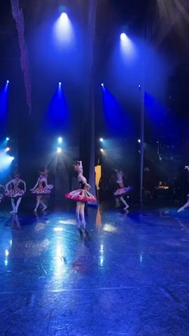 This video captures a ballet performance on a dimly lit stage. The primary subjects are several ballerinas dressed in elaborate costumes, featuring colorful, layered tutus and ornate headpieces. The lighting is dramatic, with strong blue spotlights creating a vibrant atmosphere and highlighting the dancers. The stage floor is reflective, mirroring the blue light and adding to the visual spectacle.

In the foreground, a lead ballerina is positioned centrally, her pose elegant and poised. Her tutu is a striking combination of red, white, and black geometric patterns, and she wears a detailed crown. Other ballerinas are visible in the background, some in similar costumes and others in slightly different variations. Their movements are captured in a way that suggests fluidity and grace, even as some are slightly blurred due to motion.

The setting appears to be a theatrical venue, indicated by the stage, lighting equipment, and the darkened background that suggests an audience or backstage area. The overall mood is one of artistic performance and refined entertainment. There is no visible text in the video. The time of day is not discernible, but the indoor setting and stage lighting suggest an evening performance. The location is Ufa, Russia, though this context is not explicitly visible within the video itself.