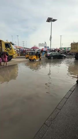 The video depicts a busy street scene in Lekki, Nigeria, with significant flooding. The foreground is dominated by a large expanse of murky water reflecting the sky, suggesting recent rain. A paved sidewalk with a raised curb is visible on the right, bordering the flooded street.

The street is populated with various vehicles and people. A large yellow dump truck is parked on the left, its wheels partially submerged. Beside it, two individuals, one wearing a pink dress, are walking. Further down the street, several yellow auto-rickshaws, a common mode of transport in the region, navigate through the water. A black car is also visible.

Numerous people are seen milling about, some under umbrellas that offer shade or protection from the elements. Market stalls or vendors are likely present, as indicated by the umbrellas and the general density of people. Streetlights with solar panels are prominent in the background, illuminating the scene. The sky is overcast, with diffused light, indicating a daytime setting, possibly late morning or afternoon.

The overall atmosphere appears to be one of everyday activity continuing despite the challenging conditions of flooding. The presence of both vehicles and pedestrians suggests a commercial or residential area experiencing normal life interrupted by water accumulation.

While most text is illegible due to distance and resolution, some markings are visible on the yellow dump truck, including what appears to be a logo or company name. The overall impression is a dynamic urban environment dealing with its unique challenges.