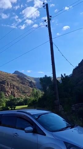 The video begins with a close-up of a silver car, specifically the front passenger side and windshield. The camera then pans upwards, revealing a bright blue sky with scattered white clouds. A wooden utility pole with power lines extends from the bottom right towards the top right corner of the frame. To the left of the pole, a steep, rocky, and arid mountain rises. The mountain is covered in sparse vegetation, with some trees visible at its base. The camera continues to pan, following the curve of the road, and the mountain's features become more pronounced. Green trees and foliage are visible in the middle ground, contrasting with the earthy tones of the mountain. The road appears to be paved and has some cracks. The sun is shining brightly, casting shadows and illuminating the landscape. The video concludes with a wider view of the mountain and the surrounding area, with the road leading into the distance. No people are visible in the video.