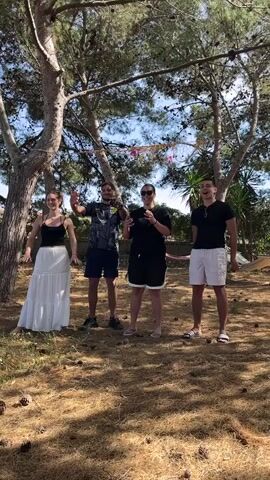 The video shows a group of four people posing for an audience seated in a clearing with trees. The "performers" are standing behind a rope strung between the trees. One is a woman with long brown hair wearing a black tank top and a long white skirt. She is positioned near the end of the rope. Next to her is a man in a dark jersey and dark blue shorts.  The third person is a woman in a black top and black shorts. The fourth person is a man in a black t-shirt and white shorts. The audience is sitting in chairs clapping and cheering at the group of four "performers." The setting is outdoors on a sunny day in a grassy, wooded area. Pine cones are strewn about the ground. Houses are visible in the background.