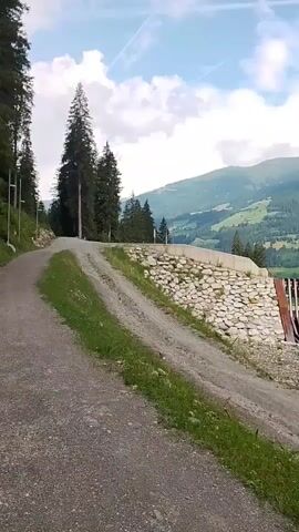 The video opens with a shot of a dirt road winding uphill, bordered by green grass and tall evergreen trees on the left. In the middle ground, a rock wall with concrete reinforcement is visible, with a structure resembling a dam or spillway to its right, featuring a series of vertical metal bars. Beyond this, a mountain range covered in greenery and trees is seen under a partly cloudy sky. The perspective then shifts, moving to the right. The camera captures a wide, shallow river with rocky banks and clear, flowing water. Dense evergreen trees line the right bank of the river, creating a thick forest. The camera continues to pan, revealing more of the river and the forest, with the mountain landscape still visible in the distance. The lighting suggests it is daytime, possibly overcast or early morning/late afternoon. No people are visible in the video.