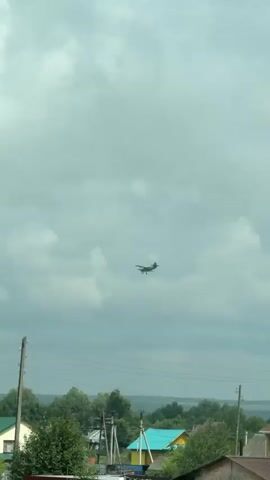 The video shows an outdoor scene from a moving vehicle. In the sky, a small propeller plane is visible against a cloudy, grey sky. Below the plane, there is a rural landscape with trees, houses with pitched roofs, and power lines. In the foreground, there are hints of green grass and foliage, suggesting a summer or late spring setting. One house in the middle ground has a yellow facade and a green roof. Another house has a brown roof. Further back, a body of water might be discernible. The video appears to be captured from a slightly elevated perspective looking upwards towards the plane.