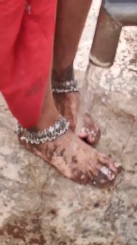 A person's legs and feet are shown as water pours onto them from a pipe. The person is wearing a red garment and ornate silver anklets and toe rings. The feet appear to be dirty before the water is applied. The ground is a rough, stone-like surface with some wetness. The video is a close-up shot focusing on the lower legs and feet.