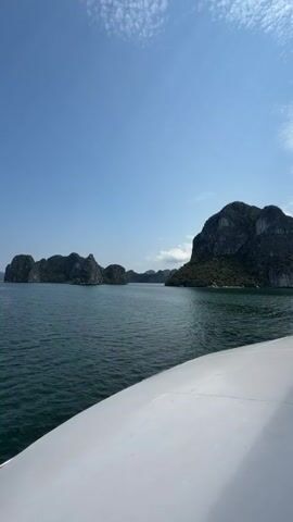 The image displays a picturesque bay scene dominated by numerous limestone karsts, characteristic of Ha Long Bay. The perspective is from a boat, with a white deck or hull visible in the lower right foreground. The water is a deep blue-green, gently rippling, and stretches towards the horizon where more islands are visible. The islands themselves are steep and rugged, covered in dark green vegetation on their upper slopes, with exposed grey rock faces. The sky above is a vibrant, clear blue with a few wispy white clouds scattered across it, indicating a bright and sunny day. There are no people visible, and no discernible text in the image. The overall impression is one of serene natural beauty, likely captured during a boat tour in Ha Long Ward, Vietnam.