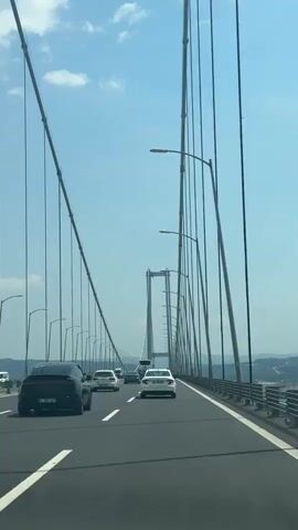The video shows a car driving on a suspension bridge on a clear, sunny day. The sky is blue with some white clouds. Other cars are visible on the bridge in both directions. The bridge has tall towers and suspension cables. The road has lane markers and guardrails. Streetlights line the edges of the bridge. Hills can be seen in the background. A person is speaking, although the speech is inaudible.
