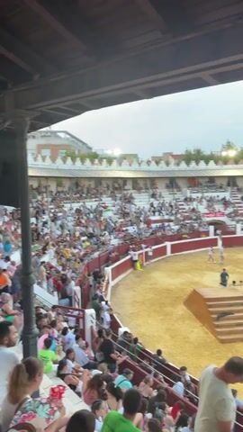 The video shows a bullring with spectators filling the stands. The arena floor is covered in sand, and there is a raised wooden platform in the center. Several people are visible in the arena, some appearing to be participants and others possibly staff. One person is standing on the raised platform. The sky is visible above the stands, suggesting it is daytime or early evening. The crowd is diverse in age and gender. The overall atmosphere appears to be that of a public event or performance.