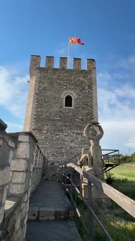 The video opens with a low-angle shot of a stone tower against a blue sky with scattered clouds. A flag, red and yellow with a sunburst, flies from the top of the tower. A stone wall with battlements runs to the left. A paved pathway leads up to the tower. To the right of the pathway, there are metal railings and a stone sculpture. The video then pans to the right, revealing more of the stone wall. The perspective shifts to a close-up of the stone wall, with the sun shining brightly. The video then cuts to a wider view from a higher vantage point, showing a cityscape with buildings, a river, a bridge, and green trees. Mountains are visible in the background. The camera continues to pan, showing more of the cityscape and a smaller stone tower with battlements in the foreground. The scene is bathed in sunlight under a blue sky.