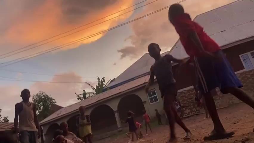 A group of children are playing in a dirt yard in front of a couple of houses. It is sunset, and the sky is colored with orange and purple clouds. The children are wearing casual clothing. The camera is tilted upwards, capturing the sky and the buildings in the background. Some of the children are running and interacting with each other.