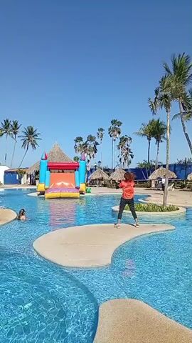 A person is exercising next to a swimming pool on a sunny day. The person has curly hair and is wearing an orange t-shirt that says "TEAM ANIMACION" on the back. They are also wearing dark pants and white sneakers. The pool is surrounded by palm trees and thatched umbrellas. There is a brightly colored inflatable slide and play area to the left of the person. The person is standing on a concrete pathway that juts into the pool and is doing arm exercises. The pool water is clear and blue. The sky is clear.