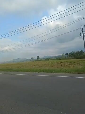 The video shows a side view of a road with a grassy field to the right. The field appears to be a harvested rice paddy with stubble. In the background, there are hills and trees under a cloudy sky with some visible power lines crossing the frame. A motorcycle with a rider wearing a helmet passes by on the road. The rider is moving from left to right across the screen. The road has a white line marking the lane. The motorcycle is black. The rider is wearing a dark shirt and jeans. The helmet is blue and black. The sky is overcast with some patches of blue.