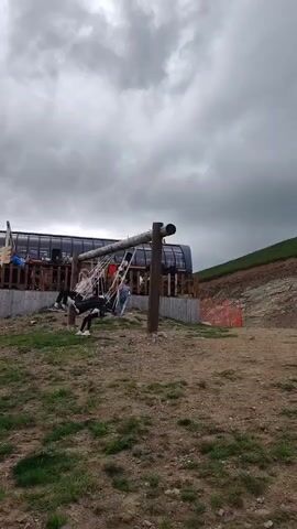 The camera pans left, revealing a mountain top with green grass and some dirt patches. A ski lift station is visible on the right, with a wooden swing set in front of it where two people are sitting. As the camera pans, more of the ski lift mechanism and support towers come into view, extending across the mountain. In the distance, other mountain peaks are visible under a cloudy sky with patches of blue. The ski lift chairs are moving, and the overall scene depicts a summer day at a ski resort.