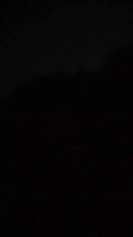 The video shows a brief flash of light against a dark background. The light appears to be purple and is illuminating the tops of trees or a tree line. The rest of the scene is extremely dark.