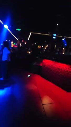 The video shows a dimly lit, busy nightclub or bar. Red lighting is prominent on a brick-like structure in the foreground and on the floor. In the background, tables are filled with people, and various lights, including white, green, and blue, illuminate the space. Some individuals are visible, mostly silhouetted or blurred. A screen displays what appears to be a cartoon character or logo in bright green. The camera moves slightly, providing a panning view of the scene.