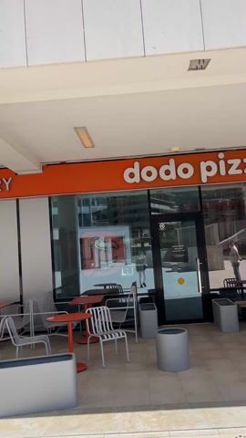 The video shows the exterior of a Dodo Pizza restaurant. The restaurant has a bright orange sign with the words "dodo pizza" in white. There is a glass door and large windows that allow a view of the interior. Several tables and chairs are set up on the patio outside. One person is visible inside the restaurant, and another walks past on the patio. The camera zooms in slightly on the Dodo Pizza sign during the video.
