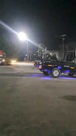 A black Mitsubishi pickup truck with blue underglow lights is parked on a paved road at night. A bright street light illuminates the area. In the distance, a white car drives past, its headlights on. The scene appears to be a street corner with buildings in the background.