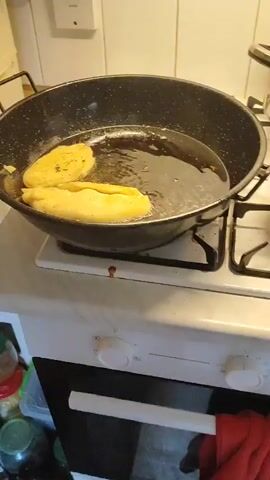 The video shows a close-up of a large, black frying pan on a stovetop. Two golden-brown pastries are frying in hot oil in the pan. The pastries appear to be crescent-shaped, with one slightly overlapping the other. The oil is bubbling vigorously around the pastries, indicating they are cooking. The pan has a black handle on the left side. The stovetop appears to be white. To the right of the pan, on the stovetop, is a white paper towel with several cooked pastries on it. These pastries are also golden-brown and vary in shape and size. In the background, a white tiled wall is visible.