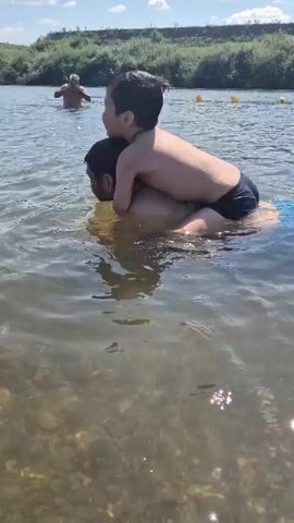 A man and a young child are in the water. The child is riding on the man's back. The man is facing away from the camera, and the child is facing towards the camera. The water is murky and appears to be a river or lake. There are yellow buoys in the background, and a hill covered in green vegetation. Another person can be seen in the background to the left, also in the water.