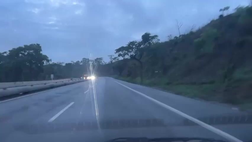 The video, shot from the dashboard of a moving vehicle, shows a first-person perspective of driving on a wet, multi-lane highway during dusk or an overcast day. The road surface is reflective from moisture. To the right, a vegetated hillside with trees runs parallel to the road. To the left, a concrete barrier or median separates opposing traffic lanes. Several vehicles, including cars and trucks, are seen in the distance approaching in the opposite direction with their headlights illuminated, casting lens flares on the windshield. The sky is a muted blue-gray, indicating low light conditions. The overall scene suggests travel on a rural or semi-rural road in wet conditions.