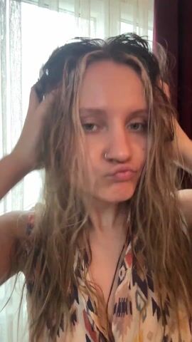 A young woman with wet, light brown hair and a nose ring is standing in front of a window with white curtains. She's wearing a patterned sleeveless top. She vigorously massages her scalp with both hands, her expression indicating discomfort or frustration. Her hair is tangled and unkempt. There's a glimpse of a red curtain behind her to the right. Music is playing in the background.