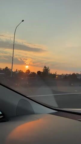 The video shows a sunset viewed from inside a vehicle. The sun is low on the horizon, casting an orange glow across the sky and clouds. Silhouetted trees and power lines are visible against the bright sky. A white car is seen driving on a road in the distance. The interior of the car, including a dashboard and window, is partially visible in the foreground. The overall mood is calm and serene.